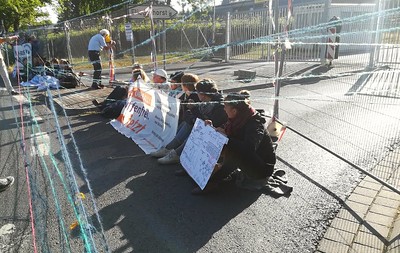 Blockade am Fliegerhorst Büchel, junge Menschen sitzen mit Schildern vor einem Zaun